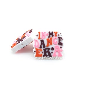 Puede incluir: Un charm de silicona blanco con el texto "IN MY DANCE ERA" en letras rosa, naranja y marrón. Las letras tienen un estilo retro.