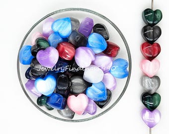 May include: A bowl filled with a variety of heart-shaped beads in colors like blue, pink, purple, and gray. The beads have a cat's eye effect, with a shimmery line running through the center. There are also a few beads strung on a wire, showing the different colors and styles.