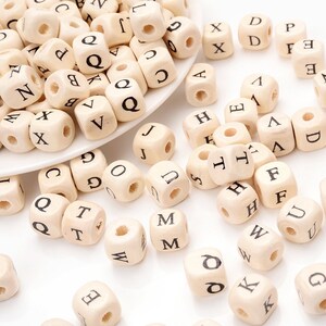 May include: A collection of small, natural wood letter beads. Each cube-shaped bead has a single black letter printed on its sides. The beads are scattered on a white surface, some in a white dish.