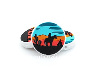 May include: A round silicone charm with a black silhouette of a cowboy riding a horse against a sunset backdrop. The sunset features orange, red, and blue colors. There are cactus plants in the foreground.