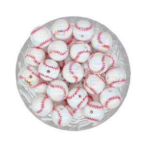 May include: A collection of white baseball-themed beads with red stitching. The beads have an iridescent sheen, and each has a hole for stringing. The beads are in a clear glass bowl.