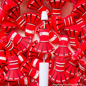 Può includere: Ciondoli e accessori a tema cheerleader rossi e bianchi. I ciondoli hanno la forma di completi da cheerleader e megafoni, con la scritta "CHEER" stampata sopra. Gli accessori includono un oggetto cilindrico bianco con un'asta di metallo e una parte superiore bianca.