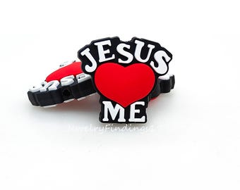 May include: A black and white shoe charm with the words "JESUS ME" in white lettering, a red heart, and a black border. The charm is on a white background.