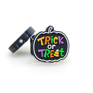 May include: Black Halloween-themed charm in the shape of a pumpkin with the words "Trick or Treat" in orange, purple, green, and white. A black charm with a silver glitter design is also visible.