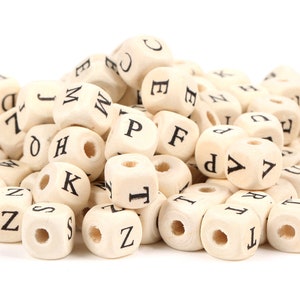 May include: A pile of wooden alphabet beads. Each cube-shaped bead is light beige with a black letter printed on each side. The beads have a hole drilled through the center, and are scattered against a white background.