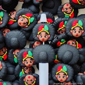 Op de afbeelding: Een verzameling zwarte kralen met afro-textuur met een cartoongezichtontwerp. Elke kraal heeft een bruin gezicht met rode lippen, een gele hoofdband en een strik met rode en groene accenten. Een wit cilindrisch object staat in het midden.