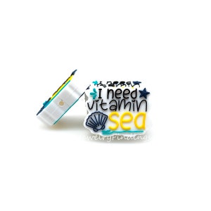 May include: White silicone charm with the text "I need vitamin sea" in black and blue lettering. The charm features a yellow seashell and a blue starfish.
