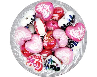 May include: A collection of heart-shaped beads in a clear glass bowl. The beads are various shades of pink and red, with white text that reads "Love You". Some beads have a swirl design.