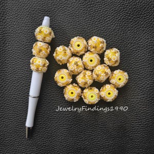 May include: A white pen adorned with daisy-themed beads in yellow and white, with black stripes. Additional daisy-themed beads are scattered nearby. The text "JewelryFindings1990" is visible. The beads are likely for jewellery making.