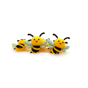 May include: Several yellow and black bee-shaped charms with smiling faces and light blue wings. The charms have black antennae and are arranged on a white background. The text "jewelryfindings1990" is visible in the bottom right corner.