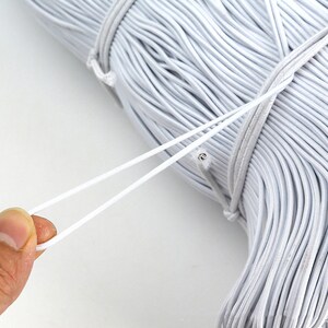 May include: A close-up of a white elastic cord wound into a coil. The cord is being pulled out of the coil, showing its elasticity.
