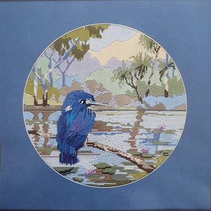 May include: A framed needlepoint picture showing a blue bird on a branch above a pond. The background features a landscape with trees, water lilies, and a soft pastel colour palette. The artwork is set against a blue mount.