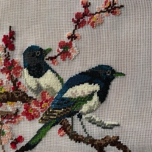 May include: Needlepoint depicting two magpies on a branch with pink and red blossoms. The birds have black, white, and blue plumage with green beaks. The background is a light beige canvas.