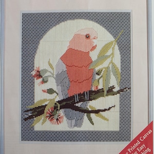 May include: Framed cross-stitch artwork depicting a pink and grey bird on a branch with green leaves and pink flowers. The bird is set against a white arched background with a grey patterned border. The artwork is framed in a silver-coloured frame.