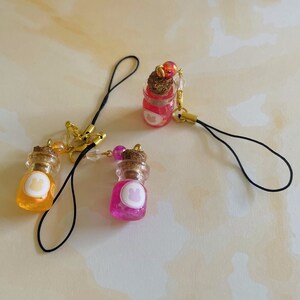 May include: Three small glass bottles with cork stoppers and colourful liquid inside. Each bottle has a white sticker with a paw print on it. The bottles are attached to black phone straps with gold accents.