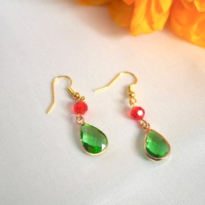 Puede incluir: Un par de pendientes dorados con una gema verde en forma de lágrima y una cuenta roja. Los pendientes, con cierre de gancho, se presentan sobre una superficie blanca. Las gemas miden aproximadamente 1,5 cm de largo.