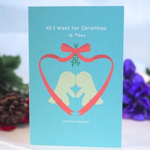 Puede incluir: Una tarjeta de Navidad azul con una cinta roja formando un corazón alrededor de dos figuras de dibujos animados verdes. El texto en la tarjeta dice "All I Want For Christmas is You" y "(and more Smaskis)".
