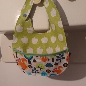 Op de afbeelding: Een groene en witte baby-slabbetje met een zakje aan de onderkant. Het slabbetje heeft een patroon van witte appels op een groene achtergrond. Het zakje heeft een witte achtergrond met een patroon van eekhoorns, bomen en paddenstoelen.