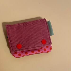 May include: A small, handmade wallet in shades of pink. The top flap is a textured, corduroy-like material, secured with two red buttons. The bottom section features a pink fabric with a red polka dot pattern. A small, green tag is attached.