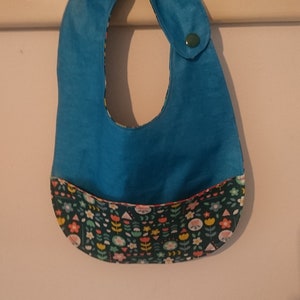 Puede incluir: Un babero azul turquesa para bebé con un bolsillo estampado con flores. El babero tiene un cierre de presión en el lateral.