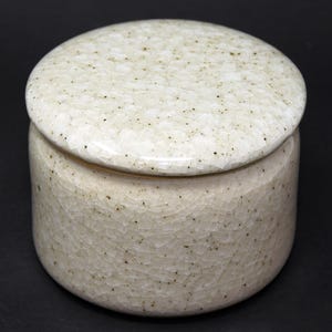 Könnte beinhalten: Ein runder, cremefarbener Keramiktopf mit passendem Deckel. Der Topf und der Deckel haben eine Craquelé-Glasur mit kleinen schwarzen Sprenkeln. Der Topf steht auf einer schwarzen Oberfläche.