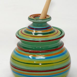 Könnte beinhalten: Ein grüner Keramik-Honigtopf mit einem Holzrührer. Der Topf ist mit bunten Streifen verziert.
