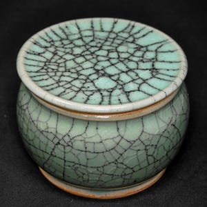 以下が含まれることがあります： 小さな丸い青磁色の陶器の壺と蓋。壺と蓋は、黒い線が入ったひび割れ模様の釉薬が施されています。蓋には白い縁があります。壺は小さく丸い台座の上に置かれています。