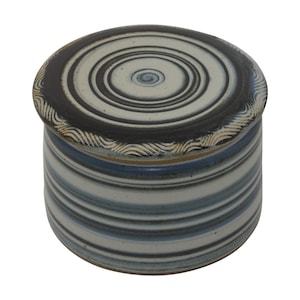 Könnte beinhalten: Eine Butterdose aus Keramik mit Deckel. Der Deckel hat ein konzentrisches Kreisdesign in Schwarz und Weiß. Die Dose ist mit blauen und weißen Streifen verziert.