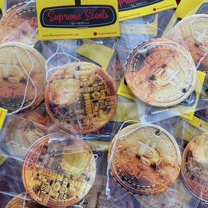 May include: A close-up of several gold-colored car air fresheners shaped like a Bitcoin coin. The air fresheners are packaged in clear plastic bags with a white string for hanging. The packaging includes the text "Supreme Scents" and "Air-Fresh-ner".