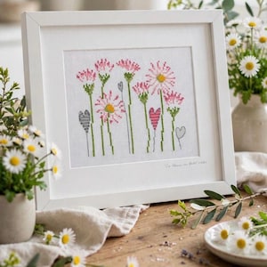 Könnte beinhalten: Eine Kreuzstichstickerei in einem weißen Rahmen mit einem botanischen Motiv aus rosa und gelben Gänseblümchen mit grünen Stielen sowie kleinen grauen und rosa herzförmigen Akzenten auf weißem Stoff, mit undeutlichem Text in der Ecke.