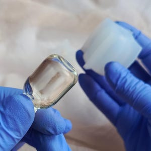 Peut inclure: Un petit flacon en verre rectangulaire transparent aux bords arrondis, présenté à côté d'un moule en silicone bleu clair translucide. Le flacon en verre présente une surface lisse et transparente, tandis que le moule a une structure creuse.