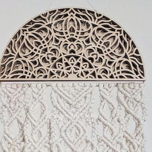 May include: A decorative wall hanging with a layered wooden design in a semi-circular shape. The intricate pattern is light brown with dark brown accents. Below, a white macrame design hangs, adding texture and visual interest.