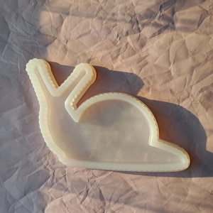 Könnte beinhalten: Eine durchscheinende, cremeweiße Ablageschale in Schneckenform mit glatter, glänzender Oberfläche und gewelltem Rand. Das Objekt hat eine hohle Mitte und zeigt die deutliche Silhouette einer Schnecke mit zwei aufrechten Fühlern aus Kunstharz.