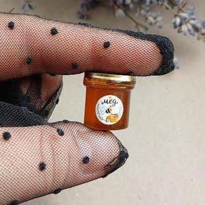 May include: A miniature glass jar filled with amber-colored honey, featuring a gold-colored metal lid and a circular white label. The label displays a small bee illustration, a honeycomb graphic, and the Cyrillic text "мед".
