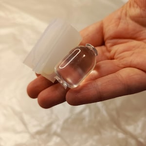 May include: A clear, smooth, miniature glass bottle with a rounded body and a narrow neck, positioned next to a translucent, flexible silicone mold shaped like a small container, both held in a palm against a white background.