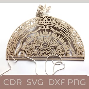 以下が含まれることがあります： 詳細なマンダラ模様が施された木製の半円形の壁掛け。壁掛けは薄い茶色の木でできており、上部に白い紐が付いています。