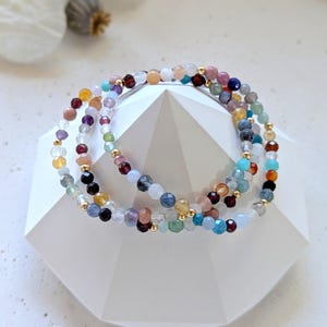 Könnte beinhalten: Ein mehrreihiges Armband mit verschiedenen bunten Edelsteinperlen, darunter blaue, grüne, rosa und violette. Das Armband besteht aus kleinen, runden Perlen und ist für einen einzigartigen Look geschichtet.