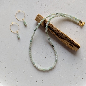 Könnte beinhalten: Ein zartes Schmuckset mit einer Halskette und einem Paar Ohrringen. Die Halskette und die Ohrringe bestehen aus kleinen, runden, hellgrünen Perlen. Die Ohrringe haben goldfarbene Reifen. Die Halskette liegt auf einem Holzstab.