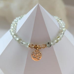 Op de afbeelding: Een armband met lichtgroene en heldere kralen en een goudkleurige boomhanger. De armband heeft een goudkleurige accentkraal en wordt gepresenteerd op een wit geometrisch oppervlak.