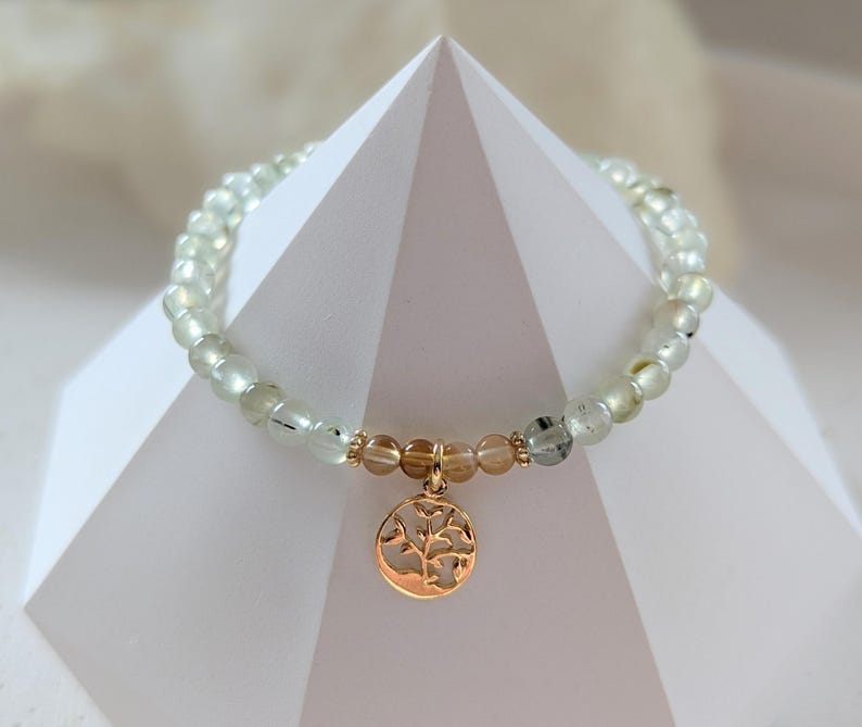 Op de afbeelding: Een armband met lichtgroene en heldere kralen en een goudkleurige boomhanger. De armband heeft een goudkleurige accentkraal en wordt gepresenteerd op een wit geometrisch oppervlak.