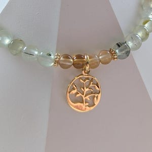 Op de afbeelding: Een kralenarmband met lichtgroene en transparante kralen. Het heeft een goudkleurige bedel met een boomontwerp. De bedel is ovaal van vorm en hangt aan een goudkleurige ketting.