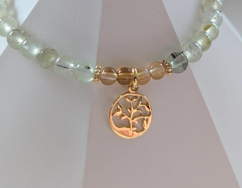 Op de afbeelding: Een kralenarmband met lichtgroene en transparante kralen. Het heeft een goudkleurige bedel met een boomontwerp. De bedel is ovaal van vorm en hangt aan een goudkleurige ketting.