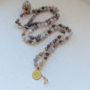 Könnte beinhalten: Eine Mala-Kette mit grauen, braunen und weißen Perlen. Die Kette hat einen goldfarbenen Anhänger mit dem Om-Symbol und einem Quastenanhänger. Die Perlen haben einen Durchmesser von etwa 8 mm. Die Mala ist für Meditation und spirituelle Praktiken konzipiert.
