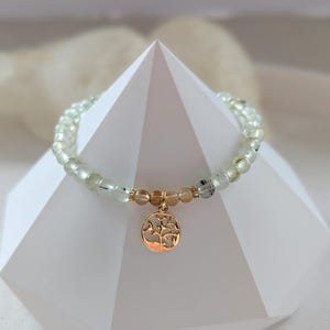 Op de afbeelding: Een armband met lichtgroene en heldere kralen, met een goudkleurige bedel in de vorm van een boom. De armband wordt getoond op een witte geometrische structuur, die het ontwerp en de kleur van de armband benadrukt.
