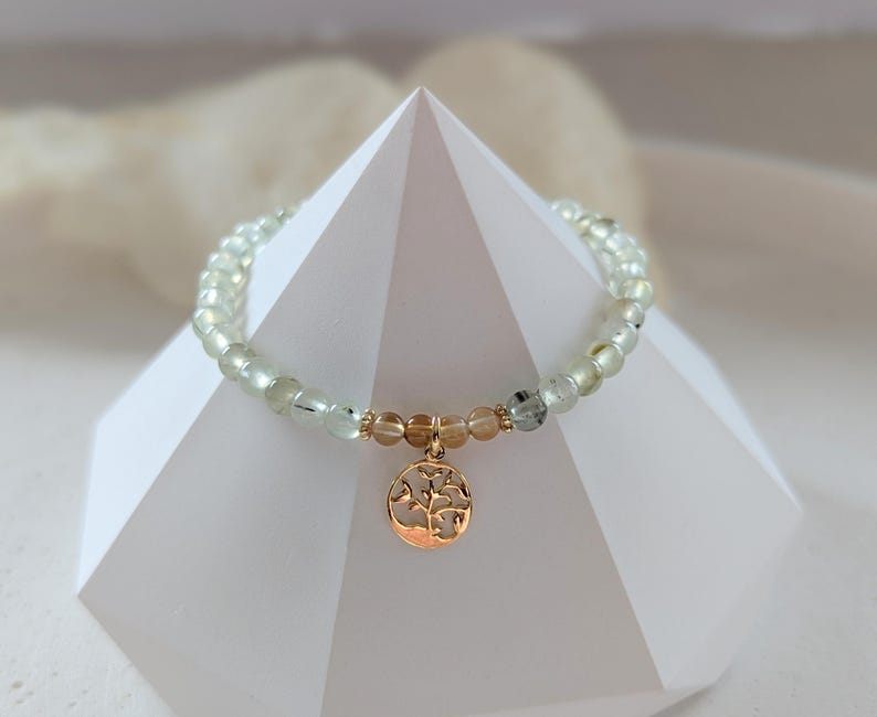 Op de afbeelding: Een armband met lichtgroene en heldere kralen, met een goudkleurige bedel in de vorm van een boom. De armband wordt getoond op een witte geometrische structuur, die het ontwerp en de kleur van de armband benadrukt.