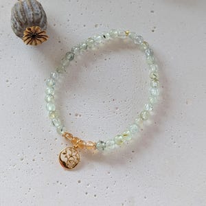 Op de afbeelding: Een delicate armband met kleine, lichtgroene kralen. Een goudkleurige bedel met een boomontwerp is bevestigd. De armband wordt getoond op een getextureerd, gebroken witte ondergrond. De kralen zijn ongeveer 0,5 cm in diameter.