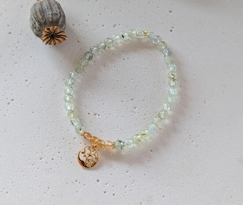 Op de afbeelding: Een delicate armband met kleine, lichtgroene kralen. Een goudkleurige bedel met een boomontwerp is bevestigd. De armband wordt getoond op een getextureerd, gebroken witte ondergrond. De kralen zijn ongeveer 0,5 cm in diameter.