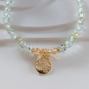Op de afbeelding: Een ketting met lichtgroene en heldere kralen. Het heeft een goudkleurige hanger met een levensboomontwerp. De hanger hangt aan een cluster van oranje getinte kralen. De ketting is een sieraad.