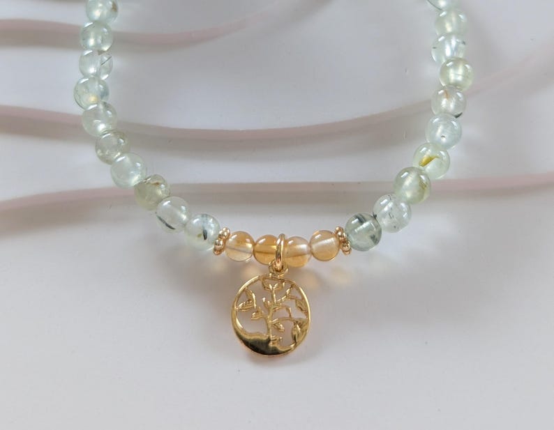 Op de afbeelding: Een ketting met lichtgroene en heldere kralen. Het heeft een goudkleurige hanger met een levensboomontwerp. De hanger hangt aan een cluster van oranje getinte kralen. De ketting is een sieraad.