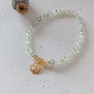 Op de afbeelding: Een delicate armband met lichtgroene en heldere kralen. Een goudkleurige bedel met een bloemmotief hangt aan de armband. De armband wordt gepresenteerd op een getextureerd, gebroken witte ondergrond. Een gedroogde zaaddoos staat op de achtergrond.